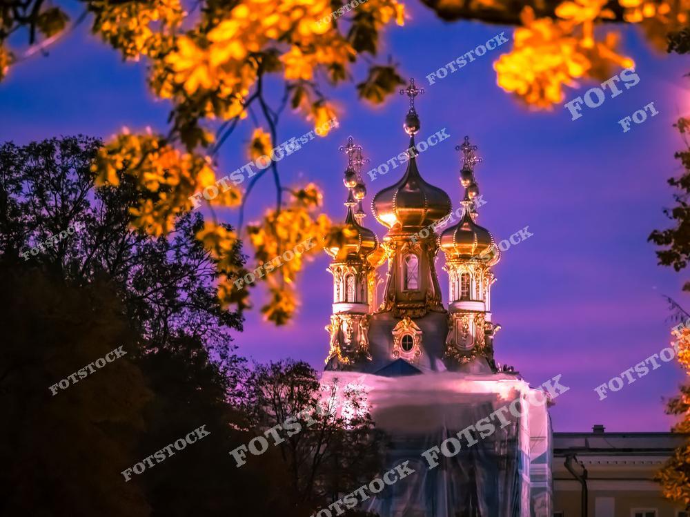 На фотографии изображена церковь Воскресения Христова Екатерининского дворца в Царском Селе. Ее золотые купола сияют в свете вечернего неба, обрамленного осенней листвой. Теплые оттенки золота и листьев контрастируют с холодными красками неба, создавая восхитительную гармонию. Этот архитектурный ансамбль – одна из главных достопримечательностей парка, привлекающая своей изысканностью.